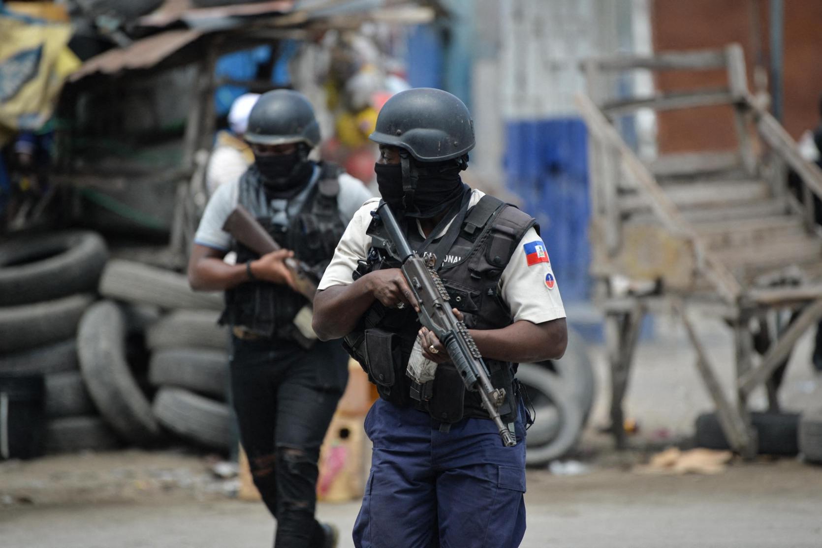 Deux agents de la Police Nationale d'Haïti (PNH), lourdement armés et portant des cagoules, progressant dans une rue parsemée de débris et de barricades de pneus.