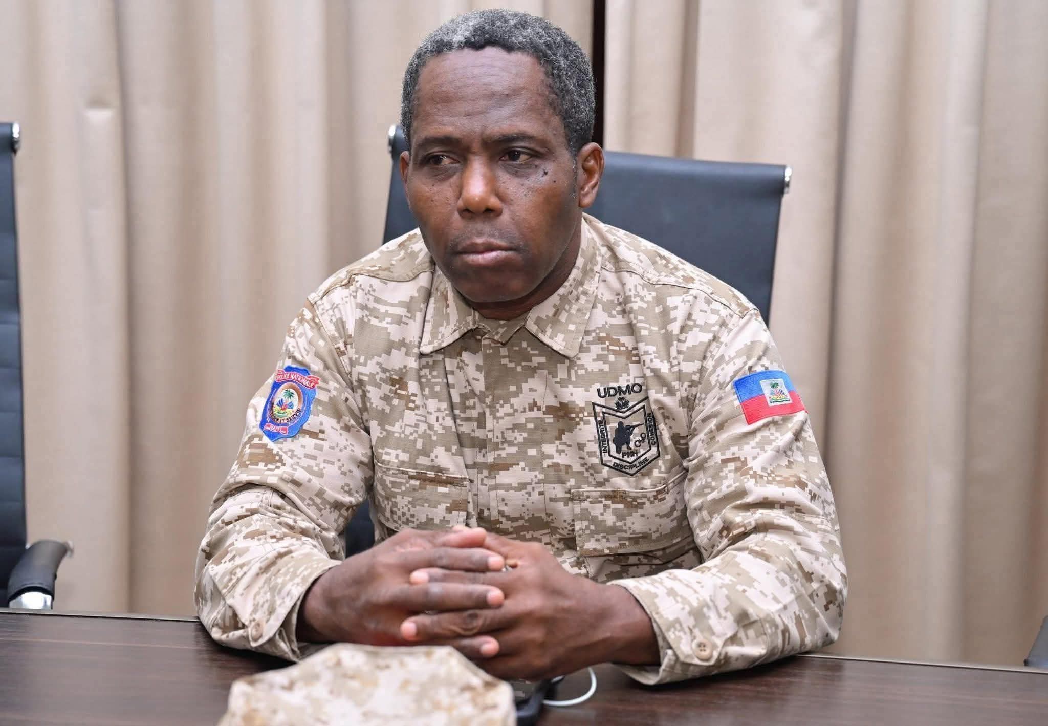 Portrait en plan moyen du commissaire divisionnaire Jacques Ader, assis à une table de réunion. Il porte l'uniforme de camouflage beige de l'UDMO (Unité Départementale de Maintien de l'Ordre) de la Police Nationale d'Haïti. Ses mains sont jointes sur la table, et l'on aperçoit les écussons de la PNH et le drapeau haïtien sur ses manches.