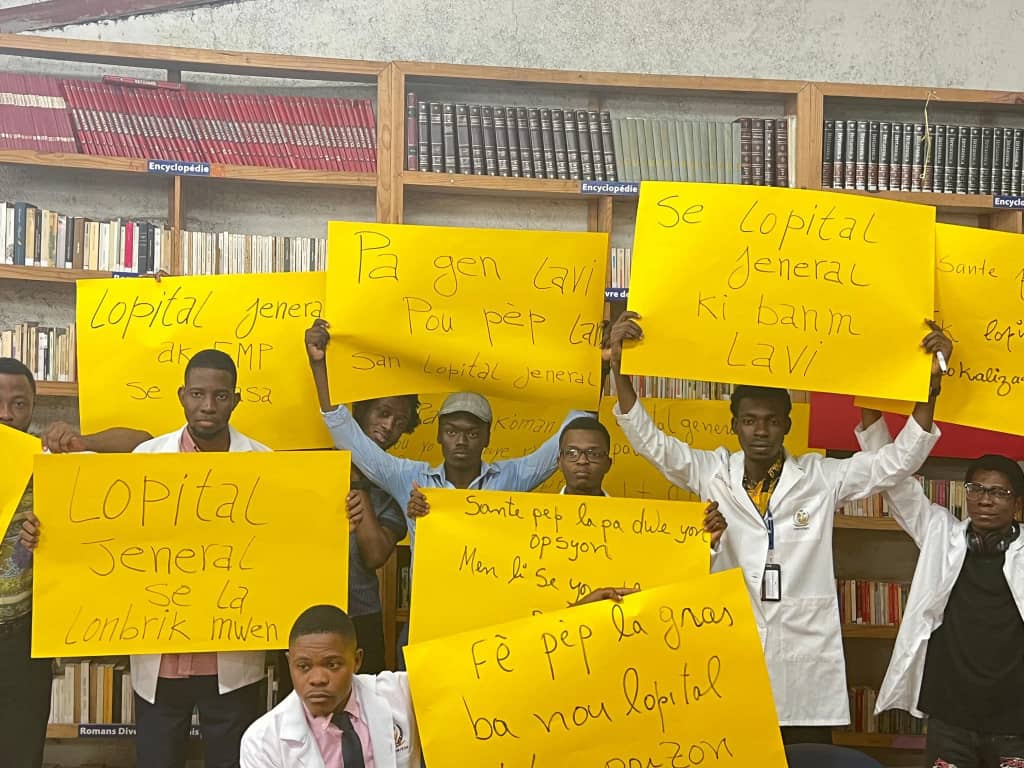 Un groupe de jeunes hommes en blouses blanches de médecine, rassemblés dans une bibliothèque, brandissant des pancartes jaunes avec des slogans en créole haïtien réclamant la réouverture et le soutien à l'Hôpital Général (HUEH).