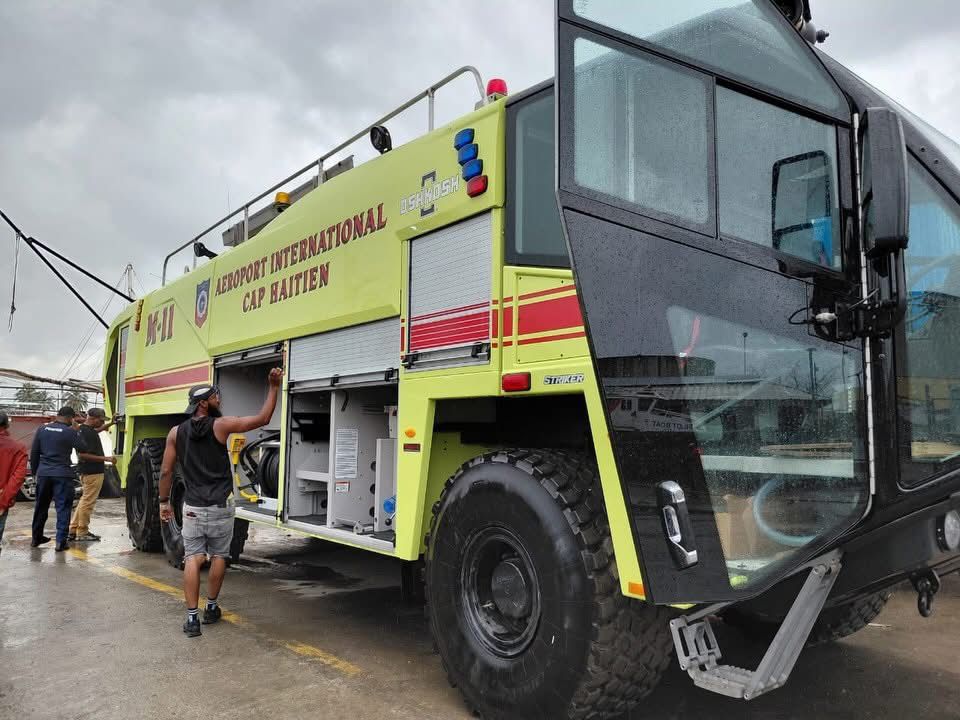 Camion de pompiers d’intervention dans les aéroports stationné à l’Aéroport International du Cap-Haïtien, avec des agents inspectant l’équipement de secours sur la piste par temps nuageux.