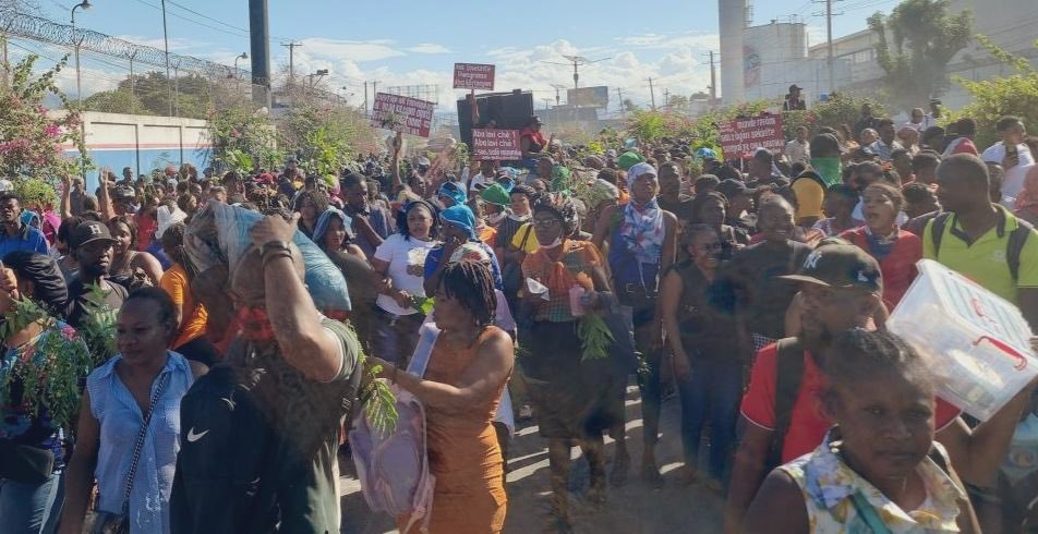Une foule compacte de manifestants marche dans une rue de Port-au-Prince. Plusieurs personnes portent des branches d'arbres en signe de protestation et brandissent des pancartes rouges avec des slogans en créole haïtien.